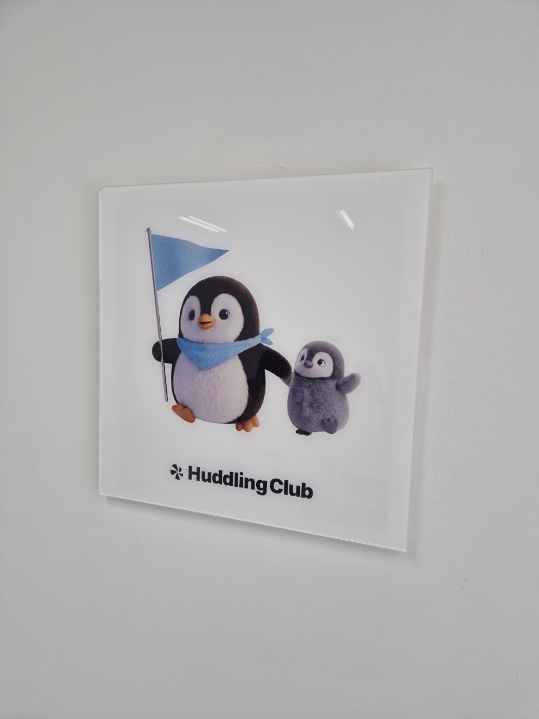 벽에 걸린 허들링 클럽 사인. 깃발을 든 펭귄과 작은 펭귄 그림 아래 Huddling Club 로고가 있다